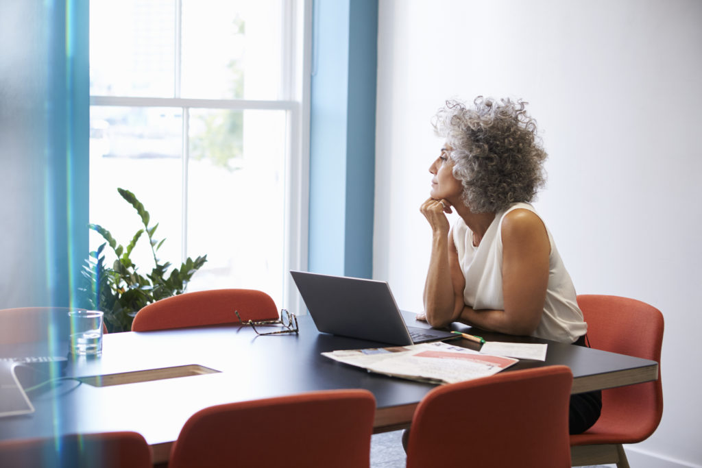 middle aged woman looking out the window while working on laptop